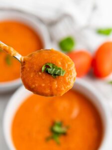 A spoonful of creamy tomato soup garnished with chopped basil, held above a bowl of the same soup.