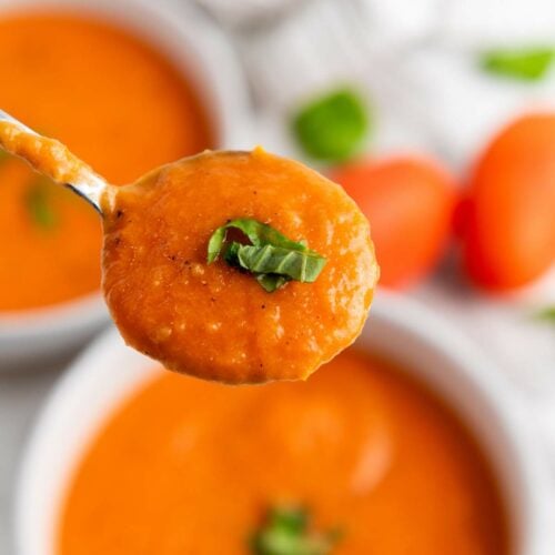 A spoonful of creamy tomato soup garnished with chopped basil, held above a bowl of the same soup.