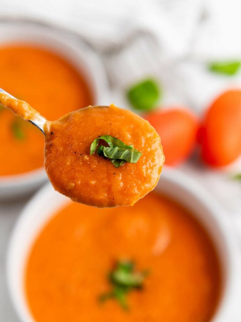 A spoonful of creamy tomato soup garnished with chopped basil, held above a bowl of the same soup.