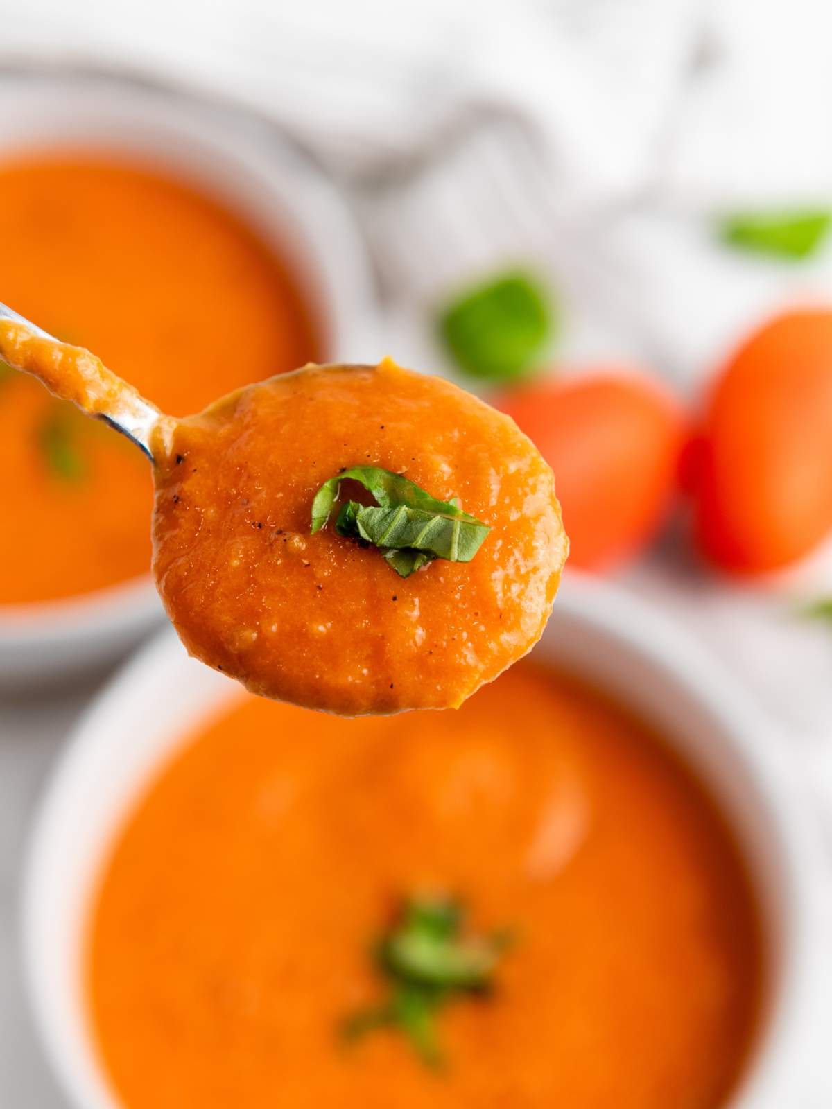 A spoonful of creamy tomato soup garnished with chopped basil, held above a bowl of the same soup.