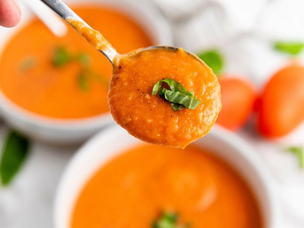 A spoonful of creamy tomato soup garnished with fresh basil held above a bowl of soup.