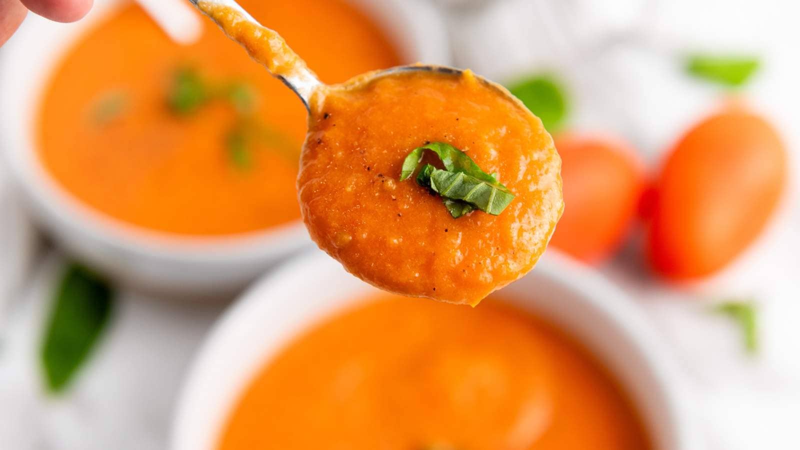 Close-up of a spoonful of tomato soup garnished with a chopped basil, with two bowls of soup and whole tomatoes in the background.