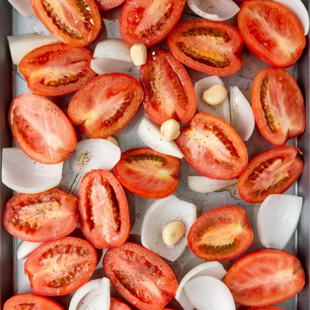 Halved tomatoes, onion pieces, and garlic cloves arranged on a baking tray, seasoned with salt and pepper.
