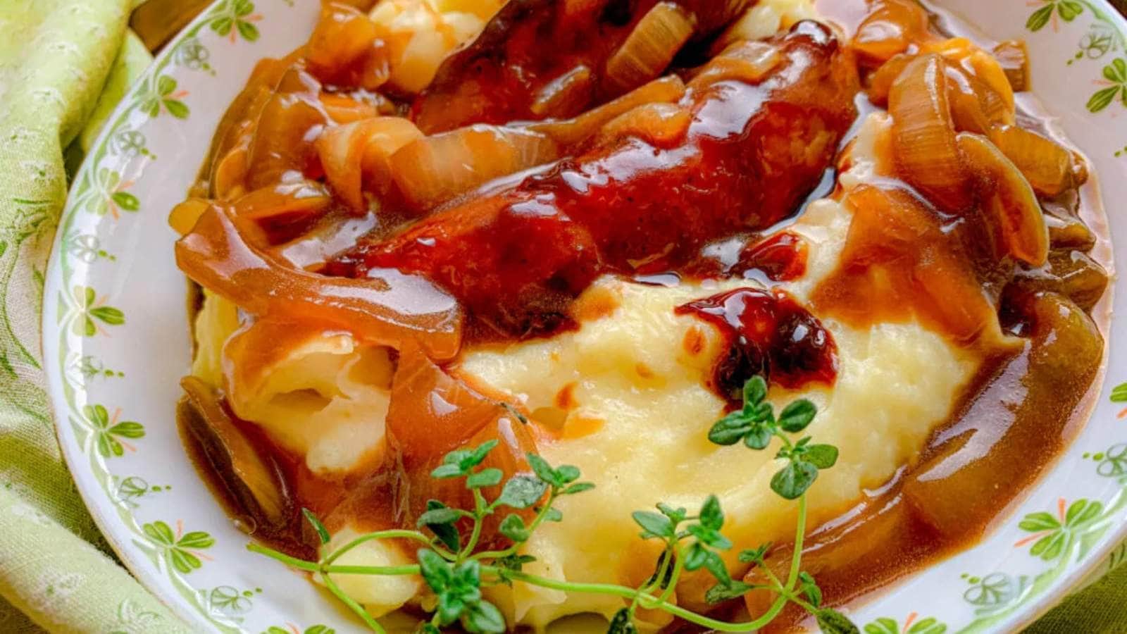 A plate of mashed potatoes topped with sausages and caramelized onions in a rich brown gravy. A sprig of fresh thyme garnishes the dish. The plate sits on a green patterned cloth.