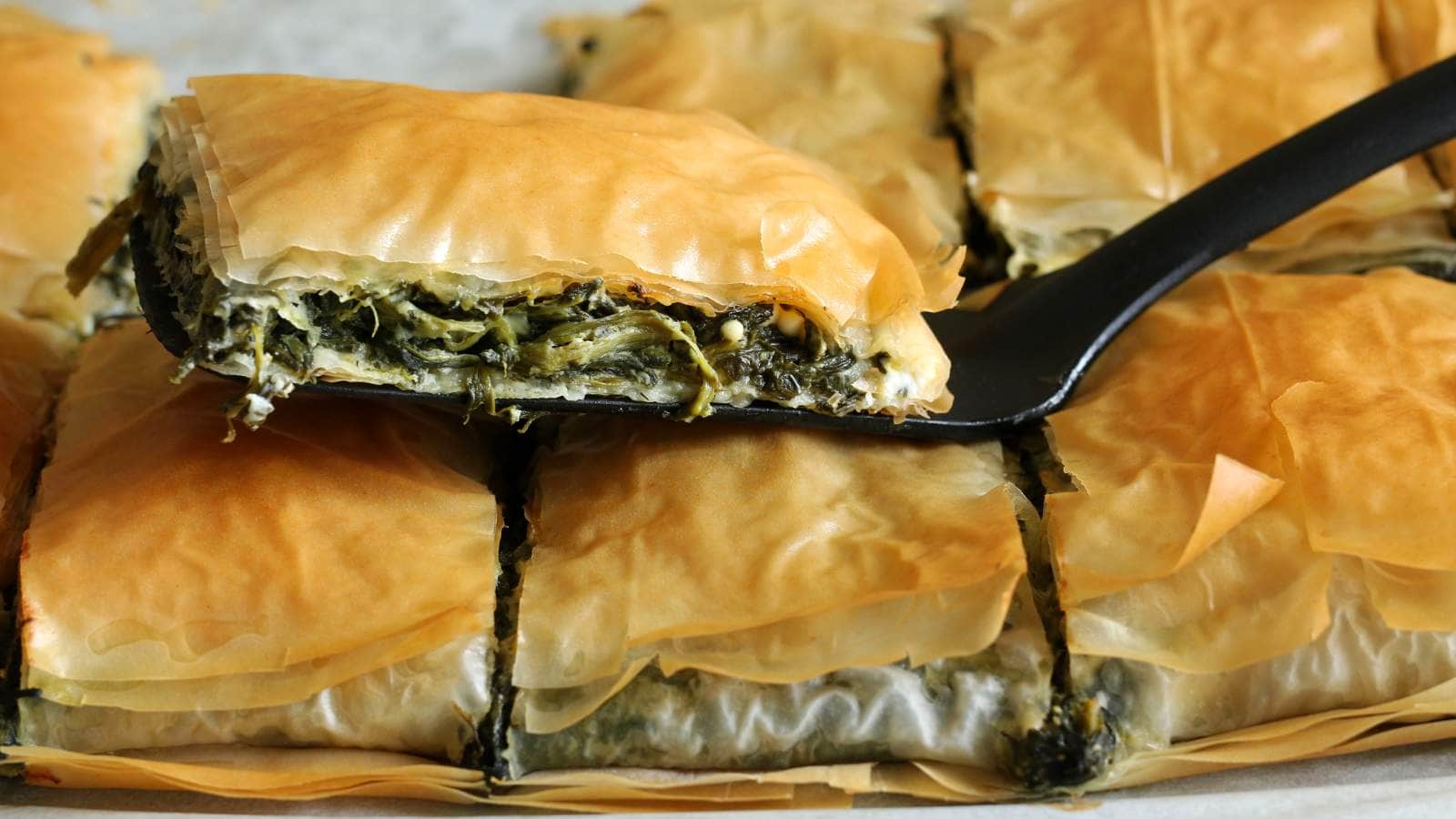 A close-up of sliced spanakopita with a piece being lifted by a black spatula, showing layers of flaky pastry and spinach filling.