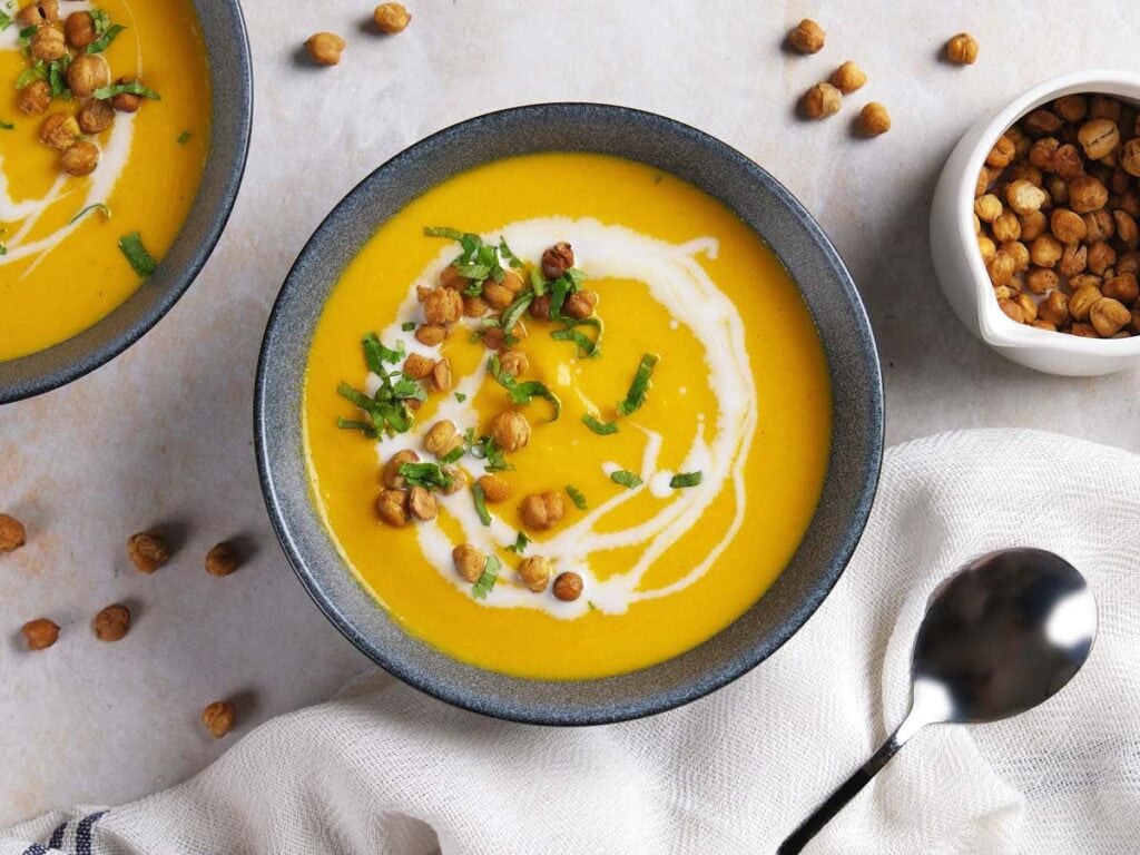 A bowl of creamy soup garnished with chickpeas, chopped herbs, and a swirl of cream, with a spoon, napkin, and a small bowl of chickpeas nearby.