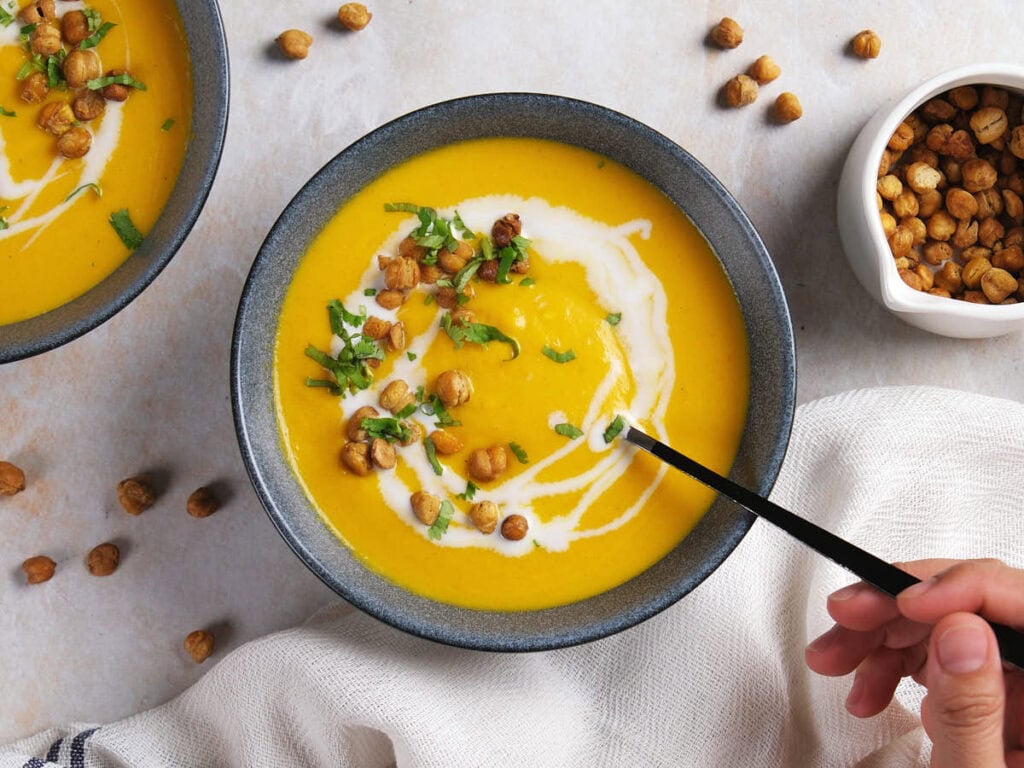A bowl of yellow soup garnished with roasted chickpeas, fresh herbs, and a swirl of cream, with a hand holding a spoon.