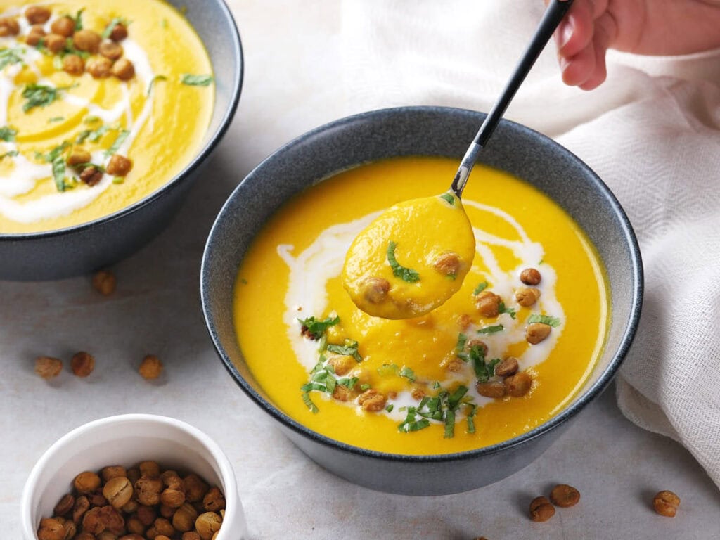 A bowl of creamy soup garnished with chickpeas, fresh herbs, and a swirl of cream.
