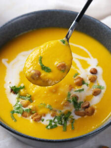 A bowl of carrot soup garnished with chickpeas, chopped herbs, and a swirl of cream, with a spoon lifting a portion.