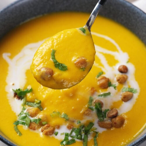 A bowl of carrot soup garnished with chickpeas, chopped herbs, and a swirl of cream, with a spoon lifting a portion.
