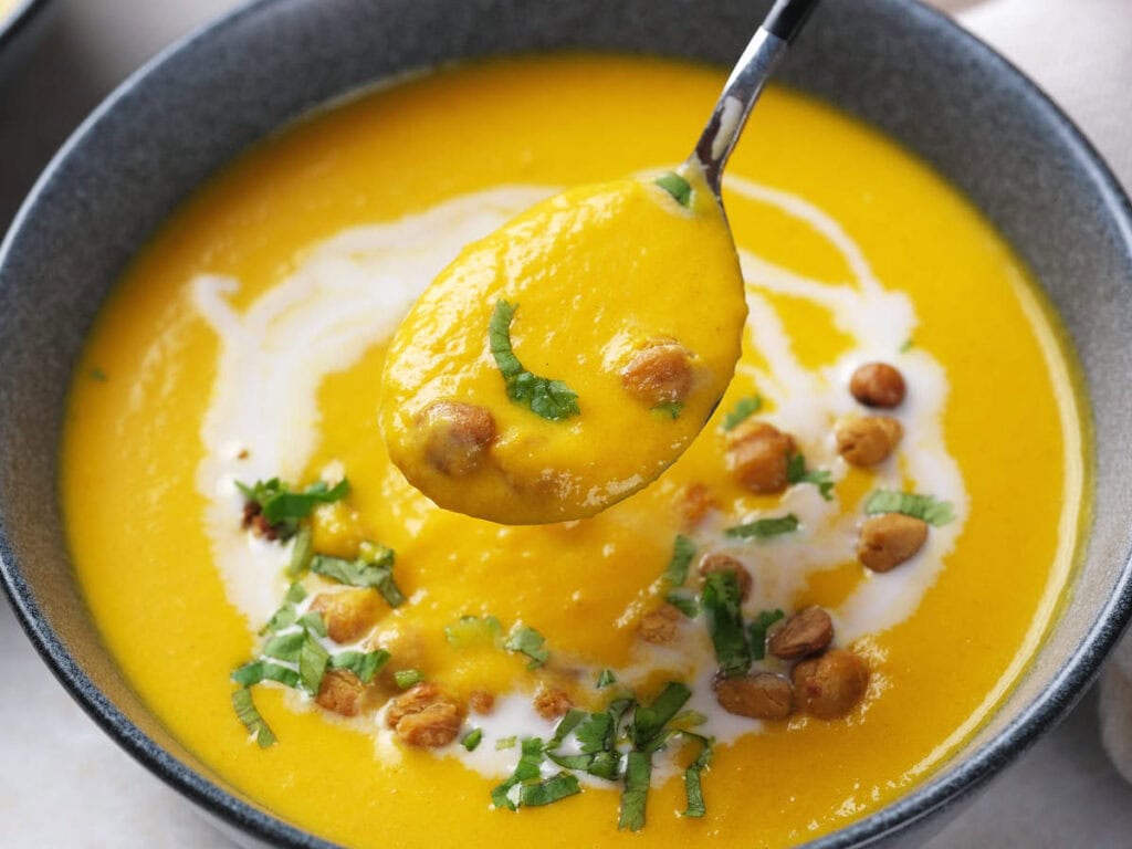 A bowl of creamy yellow soup garnished with chickpeas, fresh herbs, and a swirl of cream, with a spoonful lifted above the bowl.