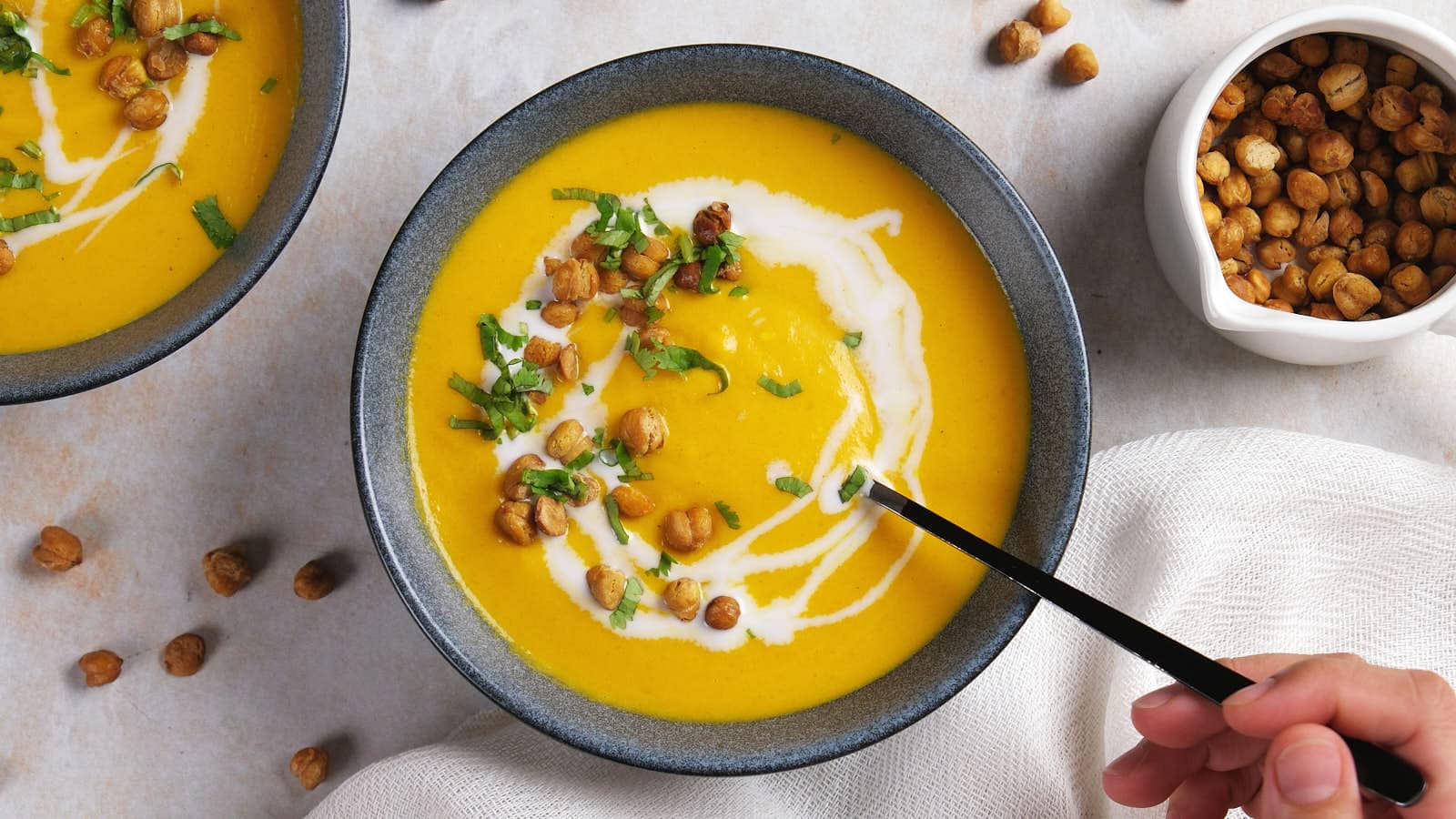 A hand holds a spoon over a bowl of carrot ginger soup topped with cream, roasted chickpeas, and herbs.