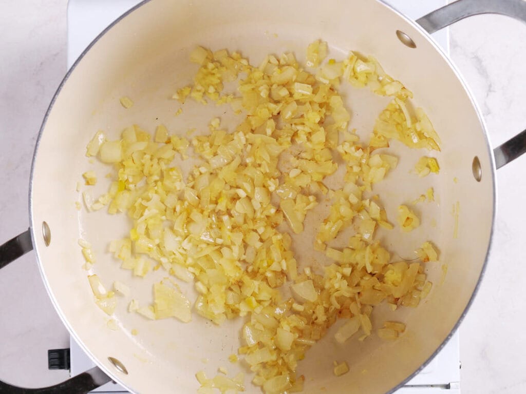 Chopped onions are sautéing in a large light-colored pan on a stovetop.