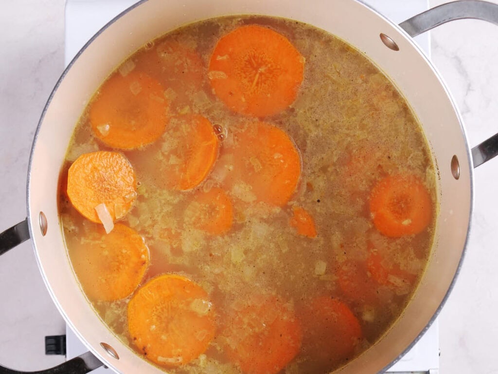 A pot of soup with sliced carrots and chopped onions simmering in broth on a stovetop.