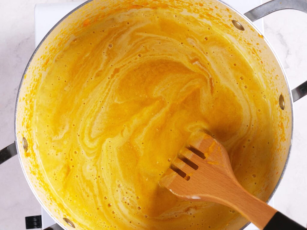 A pot of yellow-orange soup or sauce being stirred with a wooden utensil on a stovetop.