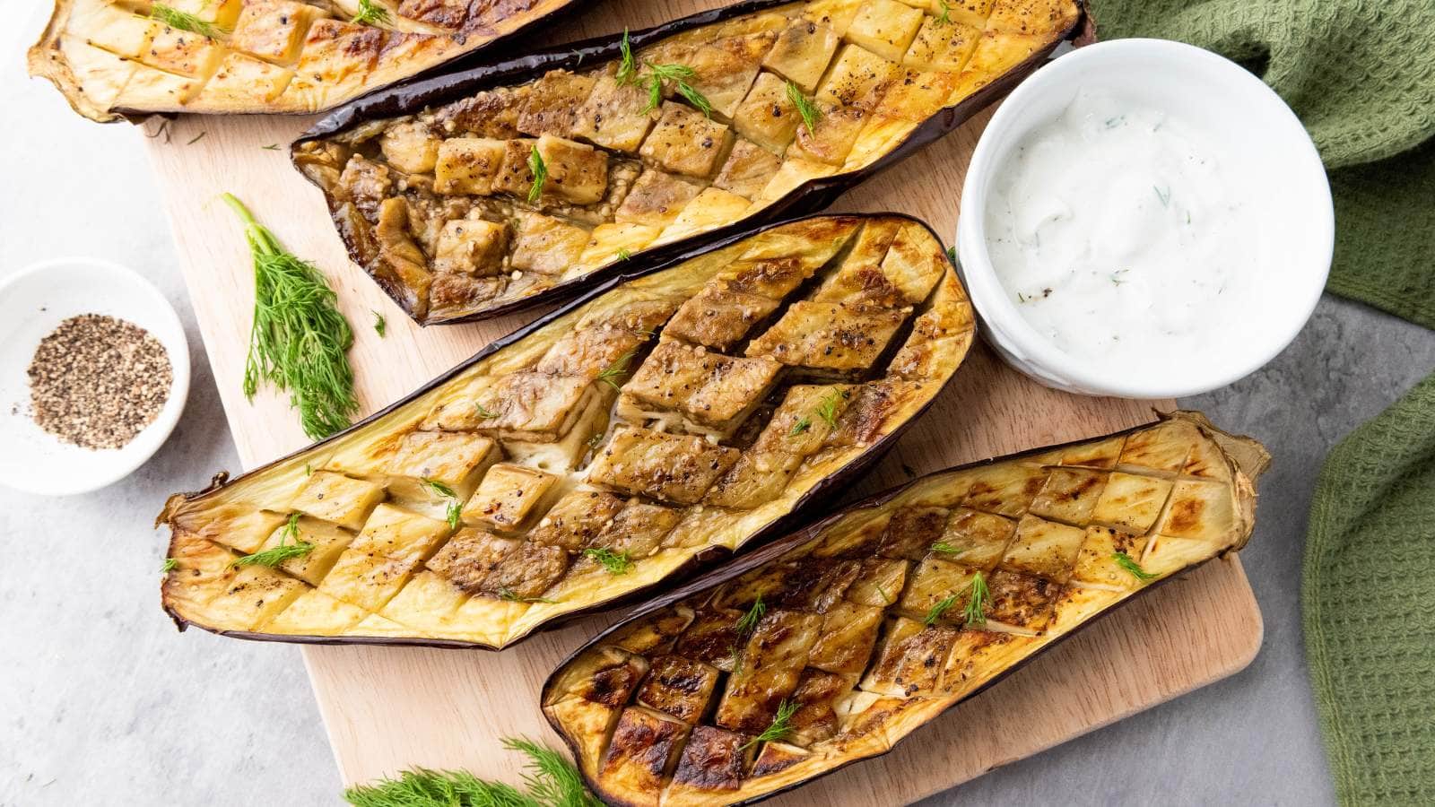 Cooked eggplant halves with a cross-hatch pattern, served on a wooden board with a bowl of creamy herb sauce.