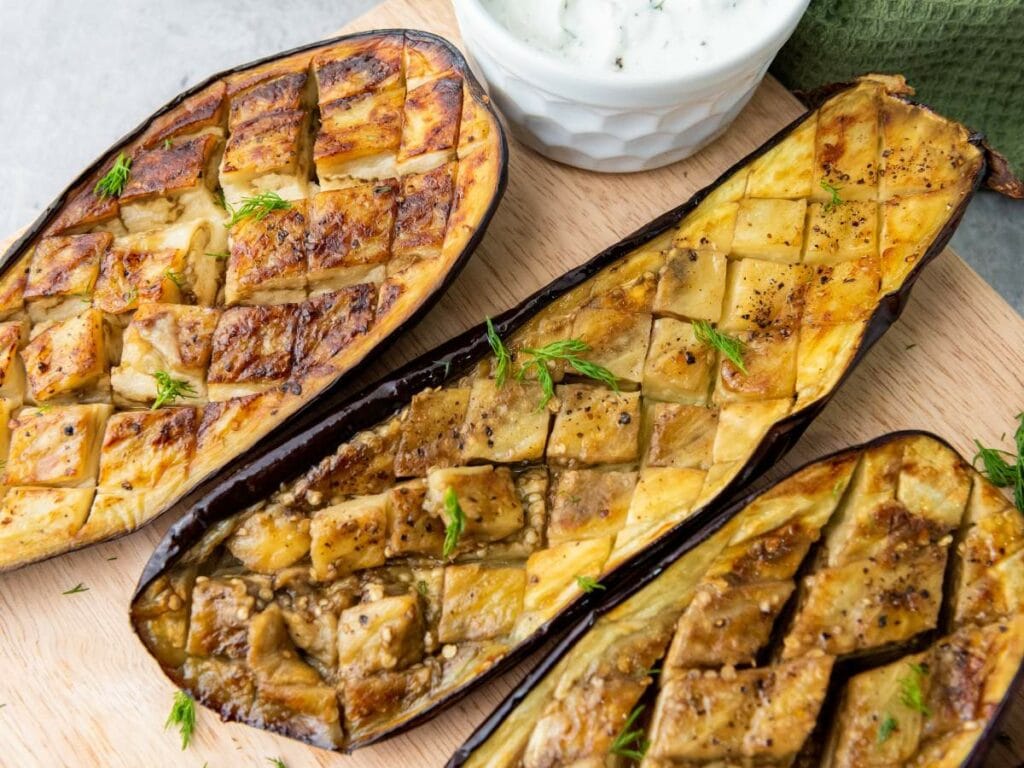 Three cooked eggplant halves with a crosshatch pattern on a wooden board, garnished with herbs.