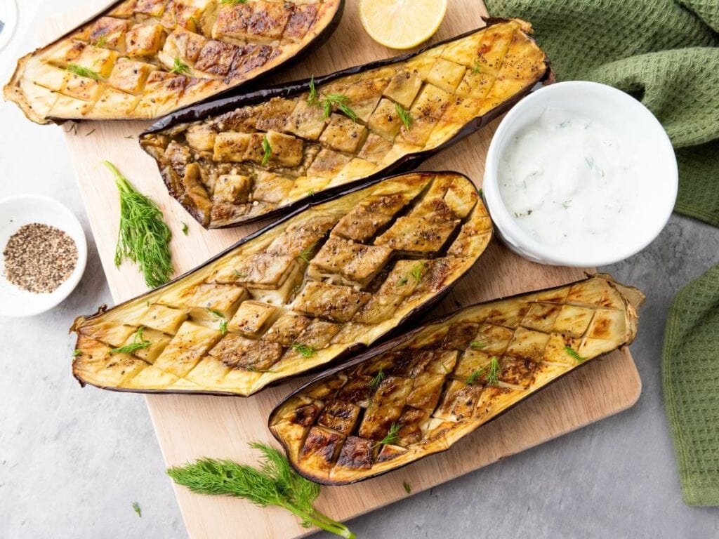 Roasted eggplant halves with a crosshatch pattern, accompanied by a bowl of yogurt sauce.