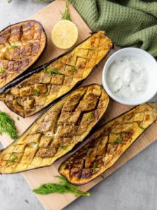 Four roasted eggplant halves with a crisscross pattern are served on a wooden board.