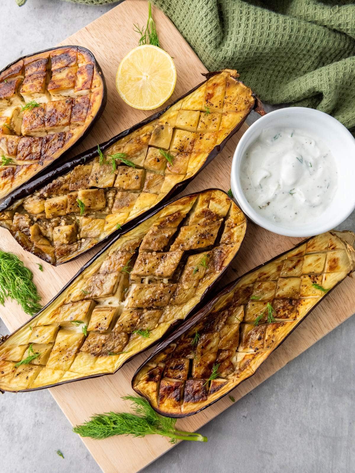 Four roasted eggplant halves with a crisscross pattern are served on a wooden board.