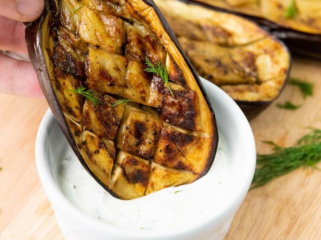A roasted, scored eggplant half being dipped into a bowl of creamy white sauce, garnished with fresh dill.