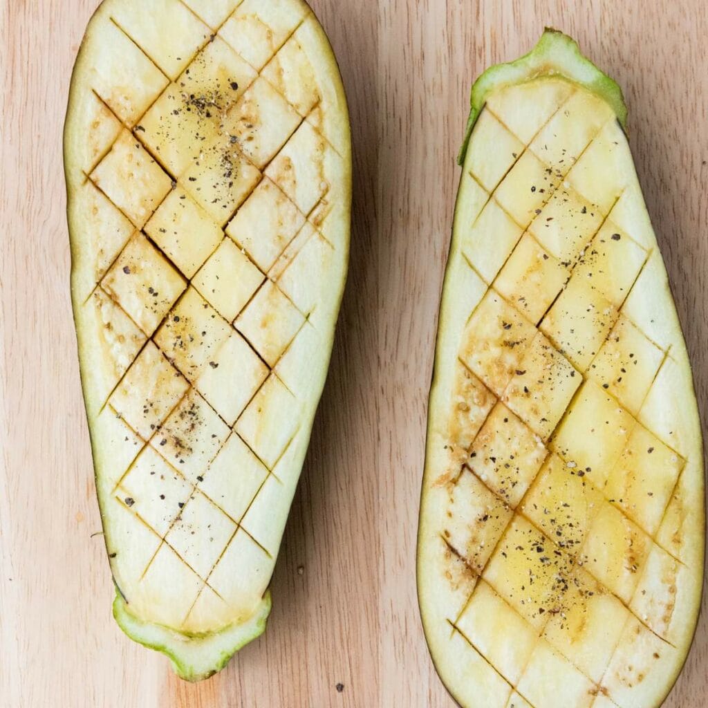 Two halves of an eggplant are scored in a diamond pattern, sprinkled with salt and pepper.