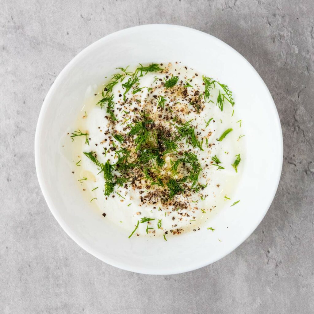 A white bowl filled with yogurt topped with chopped fresh dill, cracked black pepper, and a drizzle of olive oil.