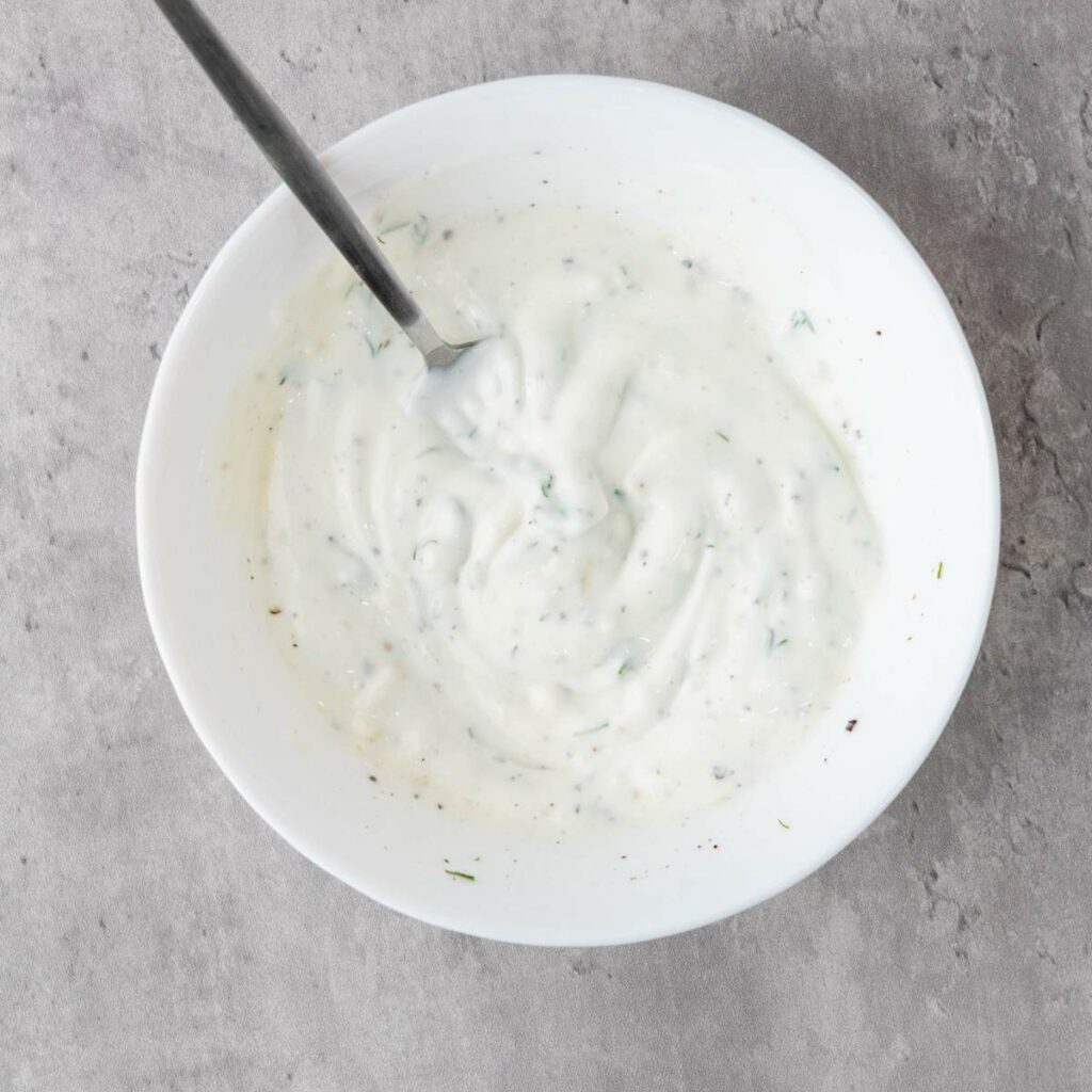 A white bowl filled with a creamy, white sauce with herbs and pepper, with a spoon resting inside.