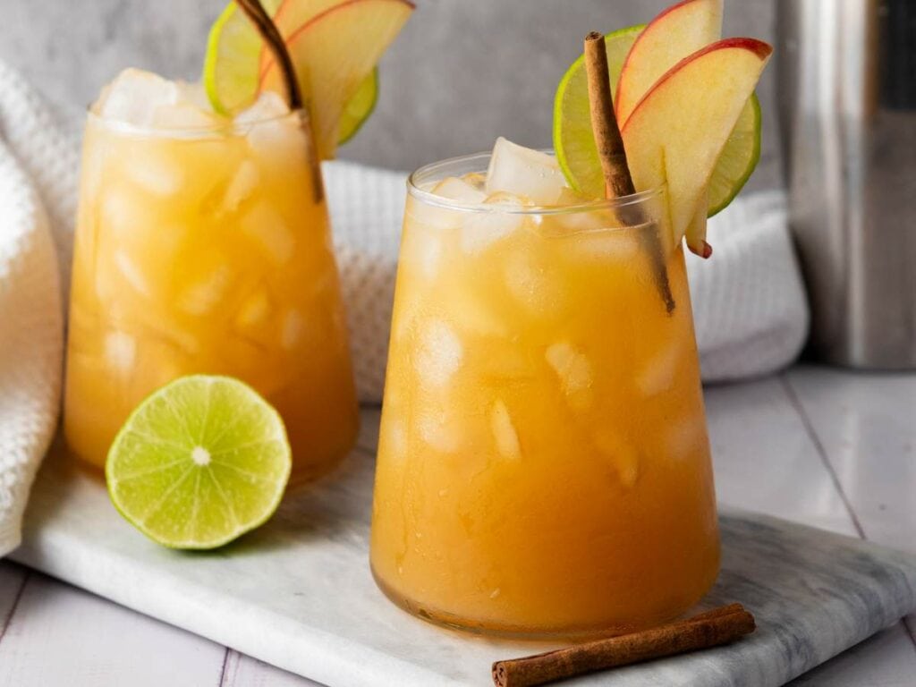 Two glasses of iced orange drink garnished with apple slices, lime slices, and cinnamon sticks.