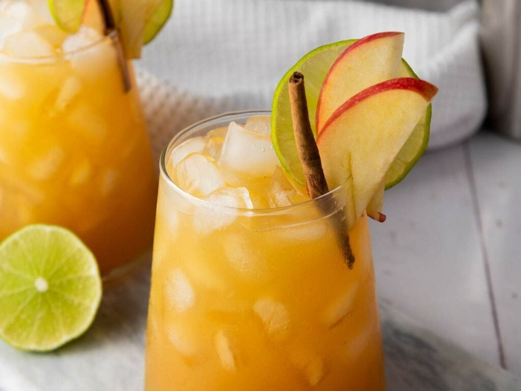 A glass of orange-colored iced drink garnished with apple slices, a lime wheel, and a cinnamon stick.
