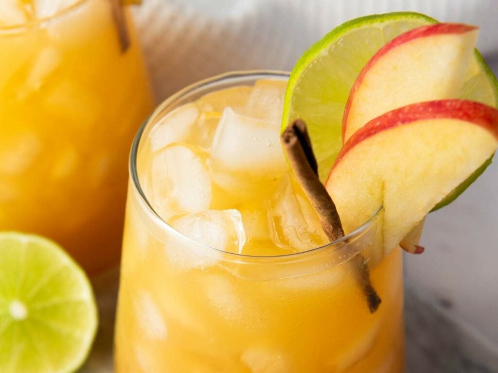 A glass of orange-colored drink filled with ice cubes, garnished with apple slices, a lime wheel, and a cinnamon stick.