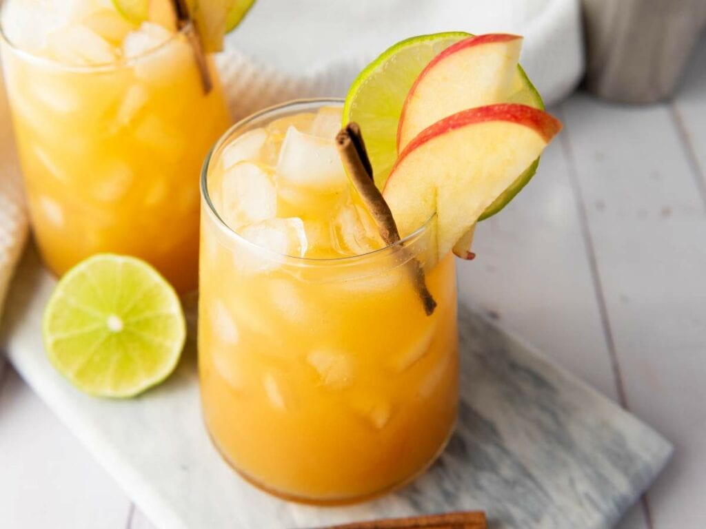 Orange iced drink garnished with lime, apple slices, and a cinnamon stick, with a halved lime and another drink in the background.