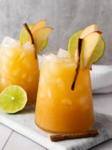 Two glasses of iced mocktail garnished with apple slices, lime slices, and cinnamon stick.