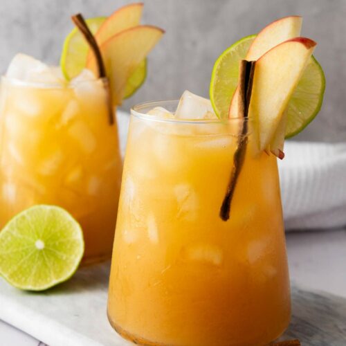 Two glasses of iced mocktail garnished with apple slices, lime slices, and cinnamon stick.