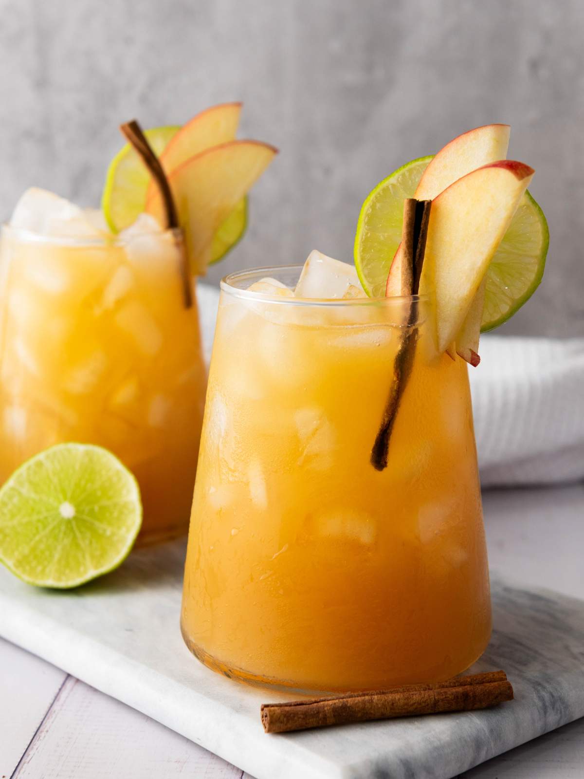 Two glasses of iced mocktail garnished with apple slices, lime slices, and cinnamon stick.