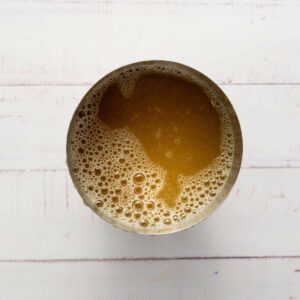 A metal cup filled with a frothy brown liquid, viewed from above, on a white wooden surface.