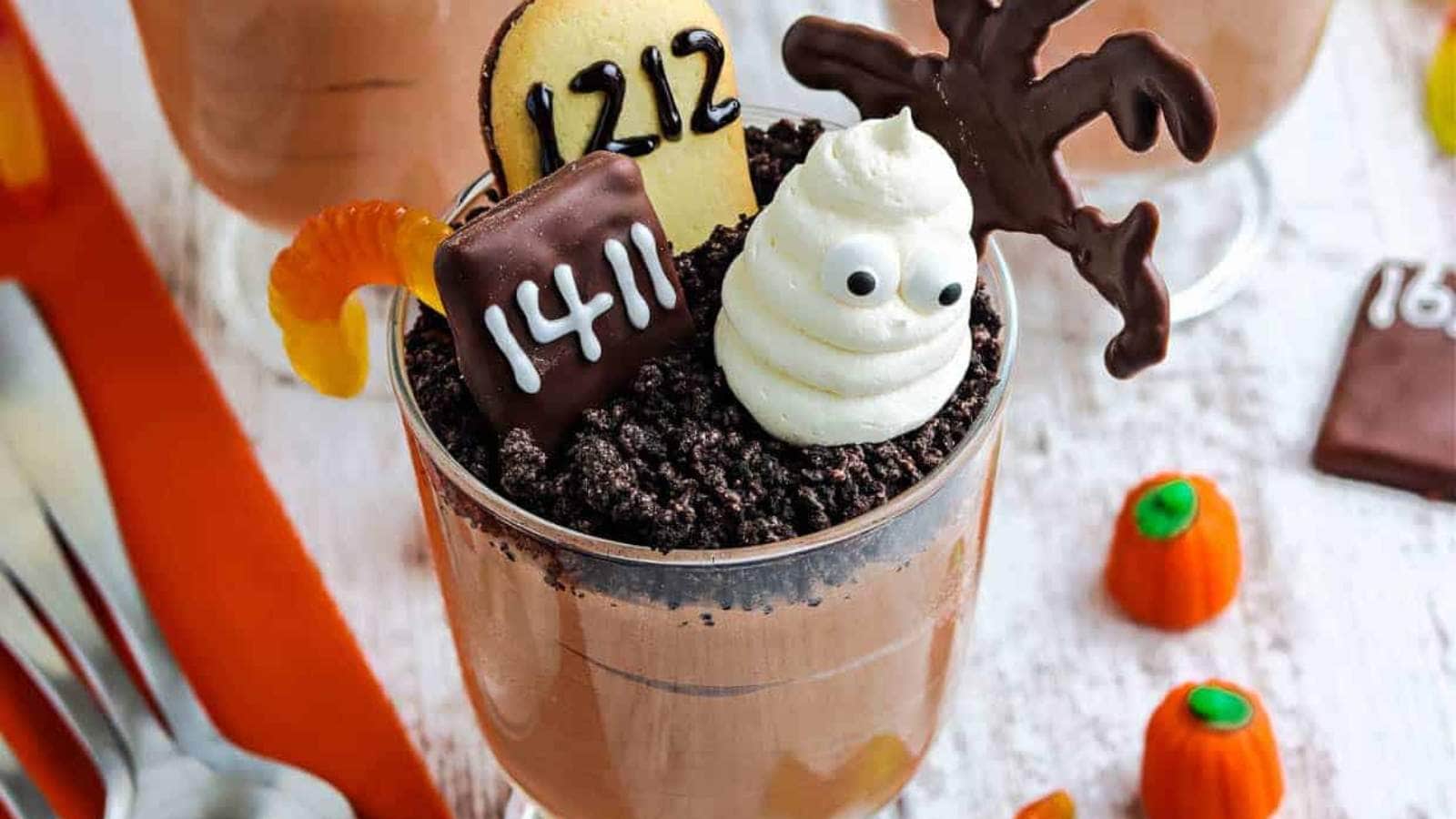 A glass cup filled with chocolate pudding, topped with cookie crumbs, a candy ghost, chocolate decorations, and candy pumpkins.
