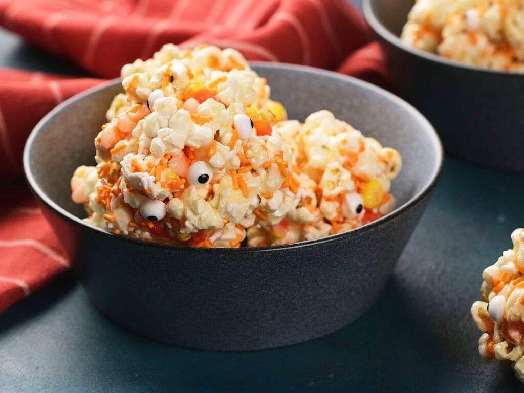 A bowl filled with popcorn balls decorated with candy corn and candy eyeballs, sitting on a dark surface.