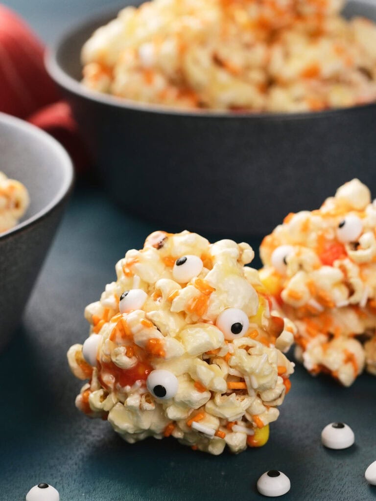 A popcorn ball decorated with candy eyes and orange sprinkles sits on a table.