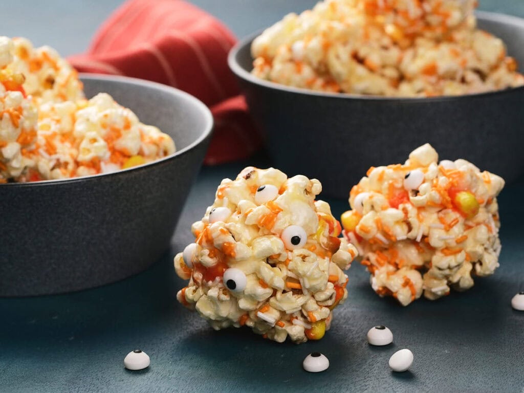Popcorn balls with candy eyes and orange sprinkles are arranged on a table, with bowls and a red cloth in the background.