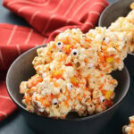 A bowl of popcorn balls mixed with candy corns and decorated with small candy eyeballs, set in front of a red striped cloth.
