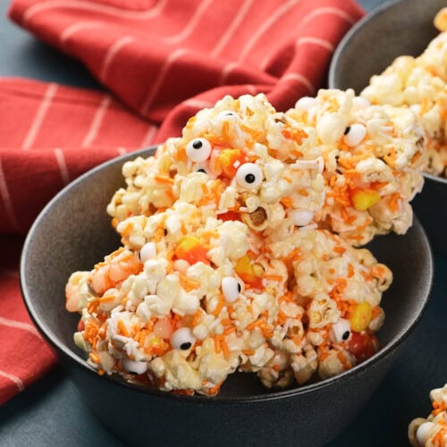 A bowl of popcorn balls mixed with candy corns and decorated with small candy eyeballs, set in front of a red striped cloth.