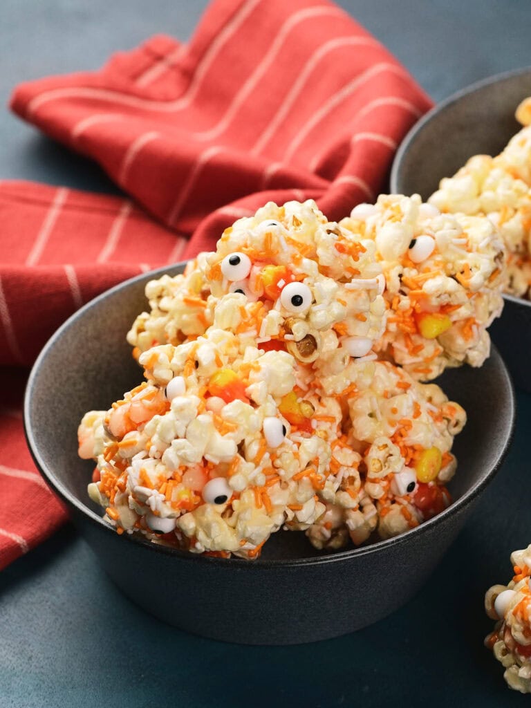 A bowl of popcorn balls mixed with candy corns and decorated with small candy eyeballs, set in front of a red striped cloth.
