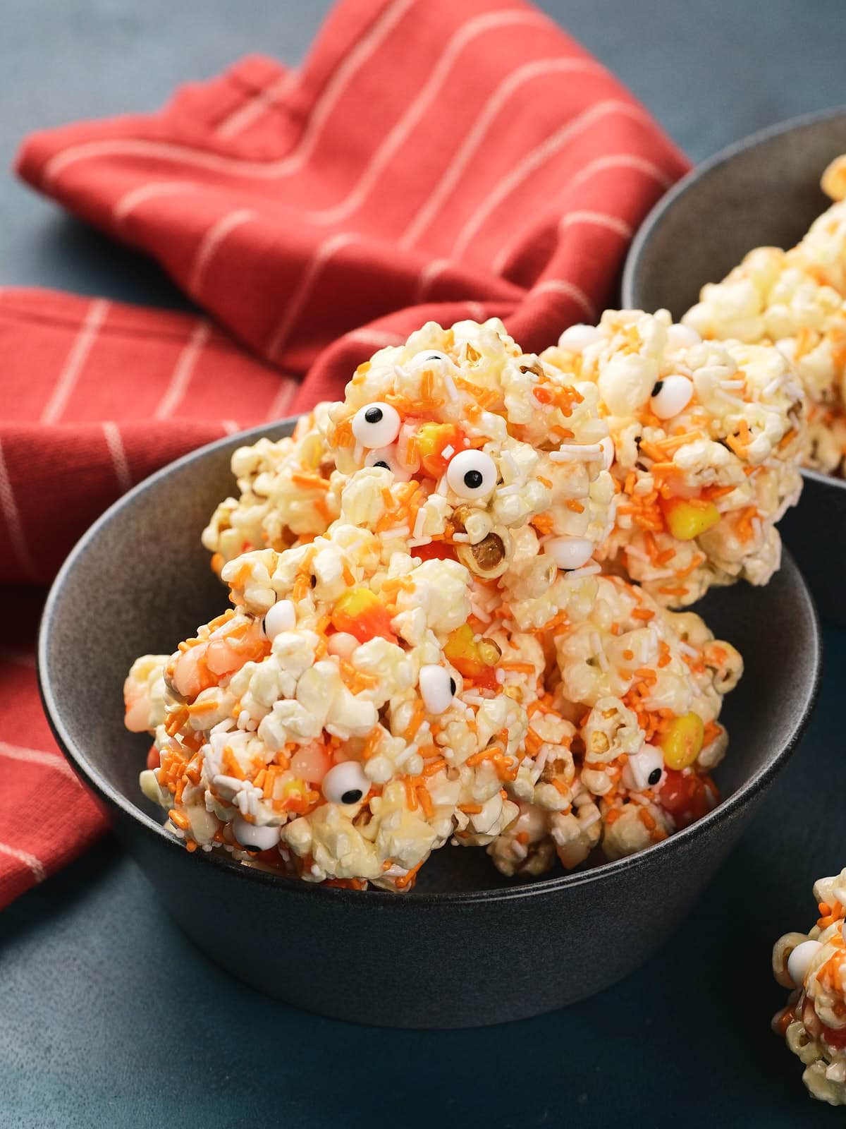 A bowl of popcorn balls mixed with candy corns and decorated with small candy eyeballs, set in front of a red striped cloth.