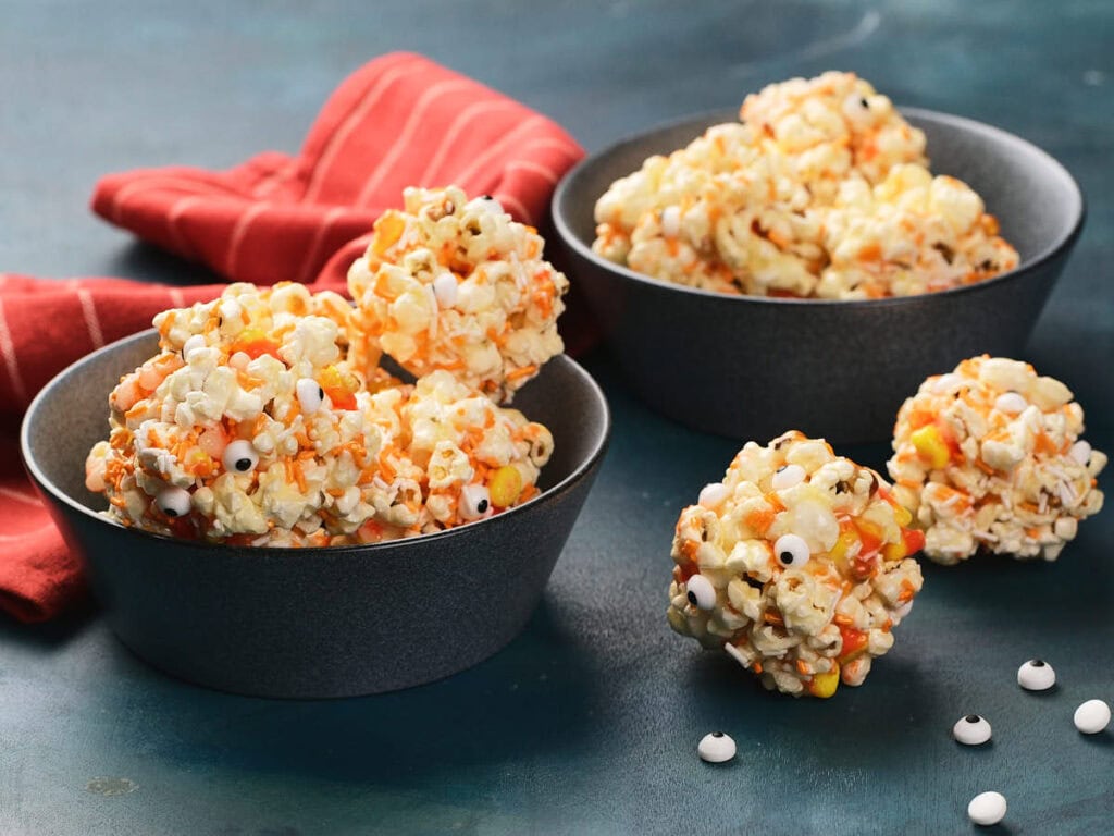 Two bowls filled with popcorn balls containing candy corn and candy eyeballs, with a red striped cloth in the background.