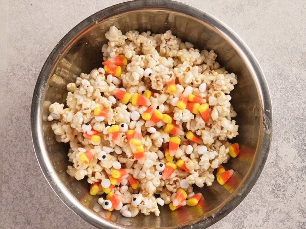 A metal bowl filled with popcorn, candy corn, small round white candies, and candy eyeballs on a light-colored surface.