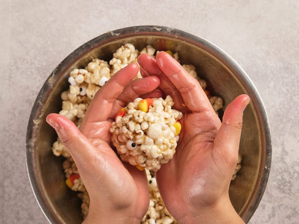 Hands shaping a popcorn ball with candy pieces over a metal bowl filled with more popcorn mixture.