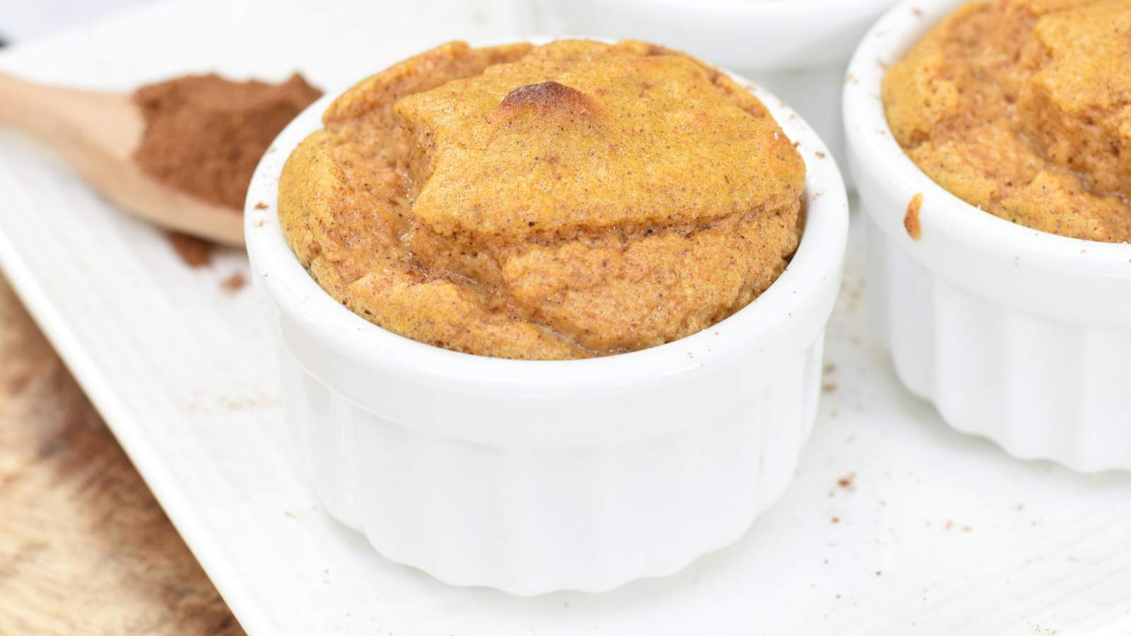 A baked soufflé in a white ramekin sits on a white plate with a wooden spoon and ground cinnamon in the background.