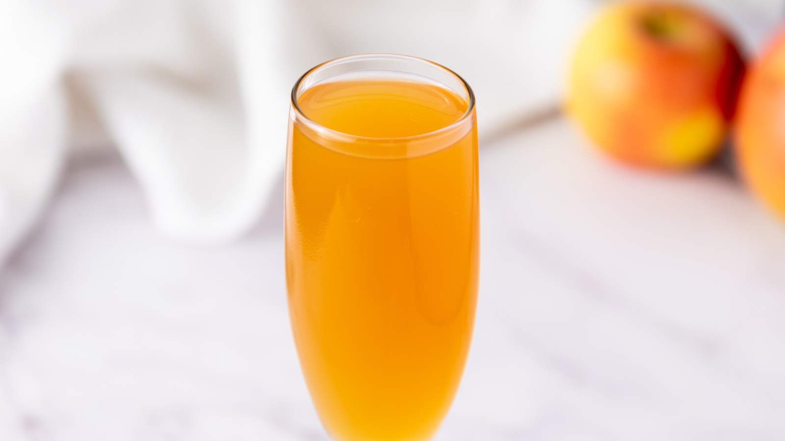 A glass of mimosa sits on a white surface with blurred apples and white cloth in the background.