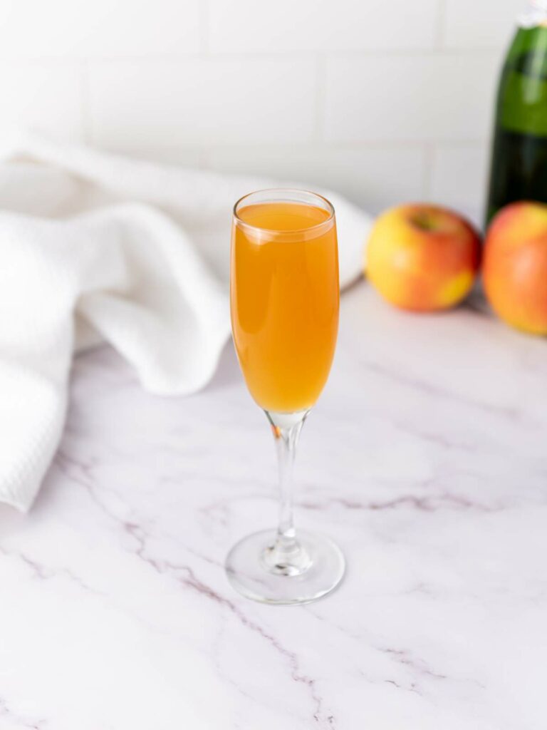 A champagne flute filled with mimosa sits on a marble surface, with apples, a white cloth, and a green bottle in the background.